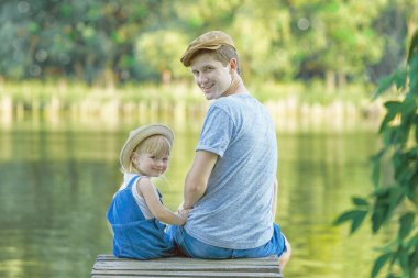 Baba ve kaba pamuklu Tulum içinde küçük bir kız köprüde oturma ve kameraya bakıyor. Arkadan görünüm, kopya alanı.