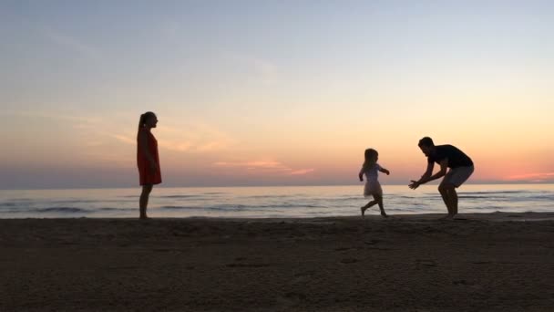 Liten flicka går mellan föräldrarna längs stranden i solnedgången. — Stockvideo