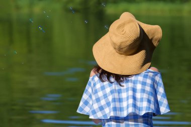 Genç kadın elbise ve kabarcıklar nehrin arka planı üfleme bir hasır şapka. Arkadan görünüm.