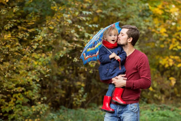 Papá tiene a una hija en las manos en un parque de otoño. Impermeable, goma — Foto de Stock