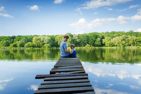 Father and a tiny girl in dungarees are sitting on the bridge an — Stock Photo, Image