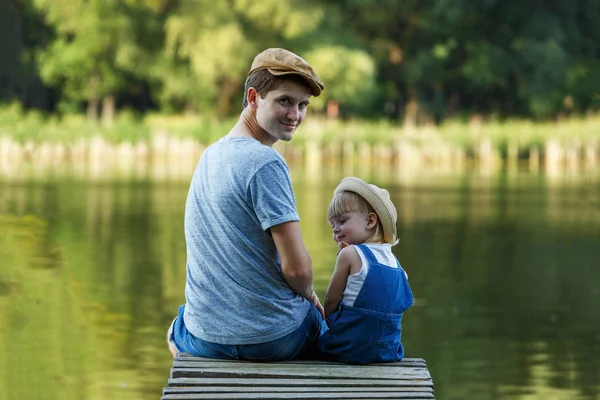 Padre y una niña pequeña en dungarees están sentados en el puente un — Foto de Stock