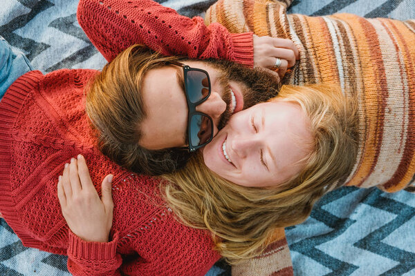 A hipster couple of blond woman and brunette man with long hair in sweaters chilling and hugging on blanket outdoors in sunrise.