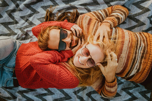 A hipster couple of blond woman and brunette man with long hair in sweaters chilling and hugging on blanket outdoors in sunrise.