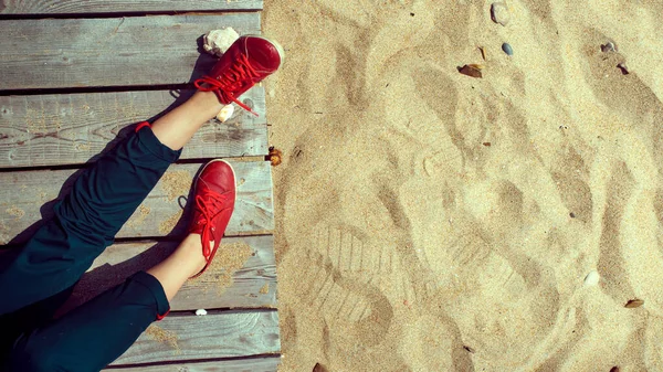 Patas de mujer en botas rojas en muelle de madera cerca de la playa de arena. Concepto de relajación, viajes y serenidad, estilo de vida — Foto de Stock