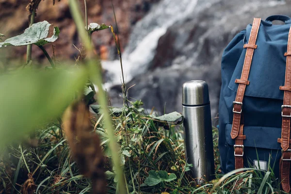 Mochila Hipster Azul, Trekking Polos y Termos Primer plano. Vista desde el bolso del viajero del turista delantero en fondo de cascada — Foto de Stock