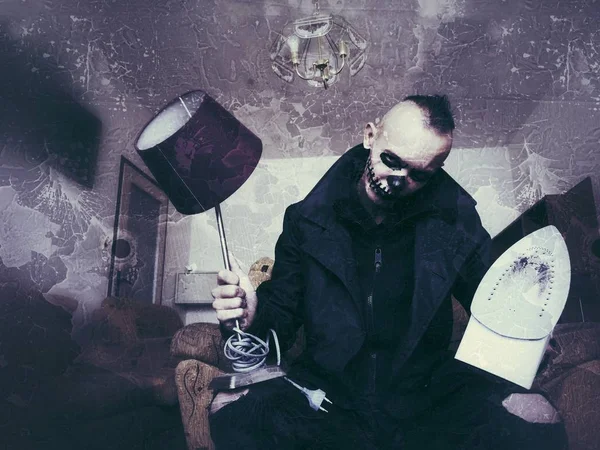 Man with scary makeup for Halloween with a table lamp and an iron in hands sitting in armchair