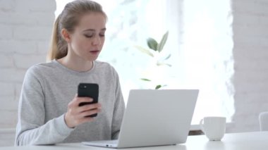 Laptop üzerinde çalışırken akıllı telefon kullanan genç kadın.