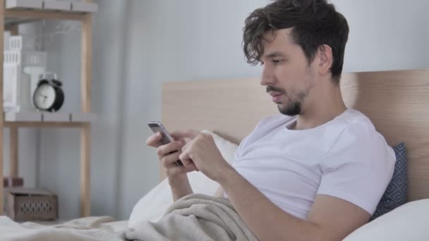 Casual Νέος Άνθρωπος Γιορτάζει Επιτυχία Ενώ Χρησιμοποιώντας Smartphone Στο Κρεβάτι — Αρχείο Βίντεο