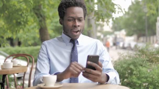 Afrikanischer Geschäftsmann bejubelt Erfolg mit Smartphone im Café — Stockvideo