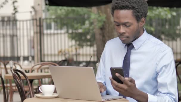 屋外カフェでスマートフォンとラップトップを使用するアフリカのビジネスマン — ストック動画