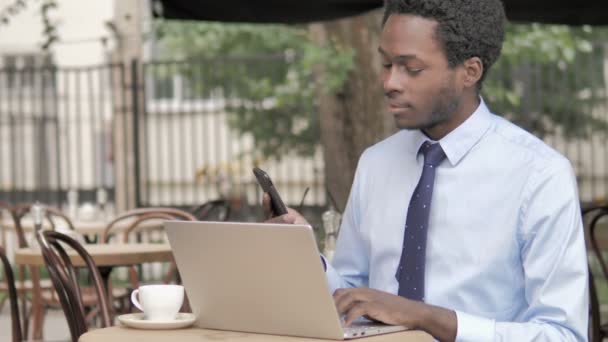 Afrikkalainen liikemies puhuu puhelimessa istuessaan ulkona kahvilassa — kuvapankkivideo