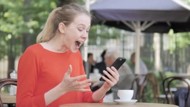 Молодая женщина аплодирует успеху на смартфоне, сидя в кафе Терраса — стоковое видео