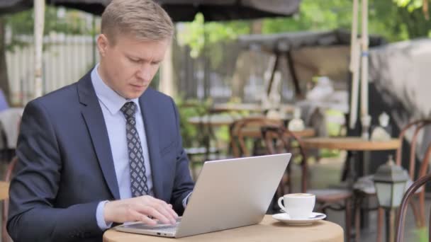 Un om de afaceri care lucrează pe laptop în timp ce stă în cafeneaua în aer liber — Videoclip de stoc