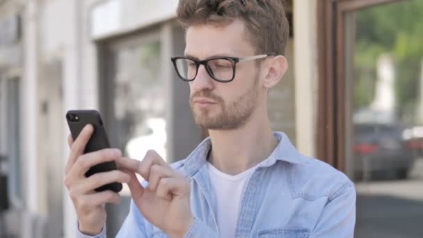 Случайный молодой человек, используя смартфон, стоя на улице — стоковое видео