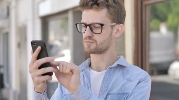 Alkalmi fiatal ember felizgat mellett elvesztés-ra smartphone rövid idő álló szabadban történő — Stock videók
