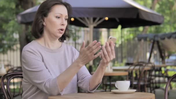 Mujer vieja molesta por la pérdida en el teléfono inteligente, sentado en el café al aire libre — Vídeos de Stock