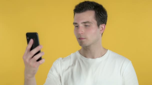 Шокований молодий чоловік використовує смартфон ізольований на жовтому тлі — стокове відео