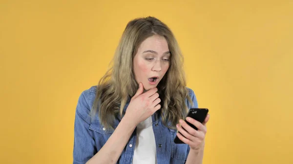 Joven chica bonita en shock mientras usa el teléfono inteligente en el fondo amarillo —  Fotos de Stock
