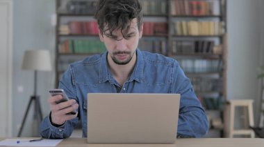 Genç adam iş yerinde akıllı telefon ve dizüstü bilgisayar kullanıyor.