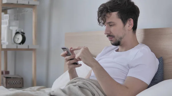 男子使用智能手机, 而躺在一边在床上 — 图库照片