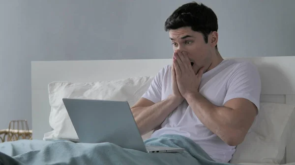 Mladý muž rozrušený ztrátou při práci na přenosném počítači v posteli — Stock fotografie