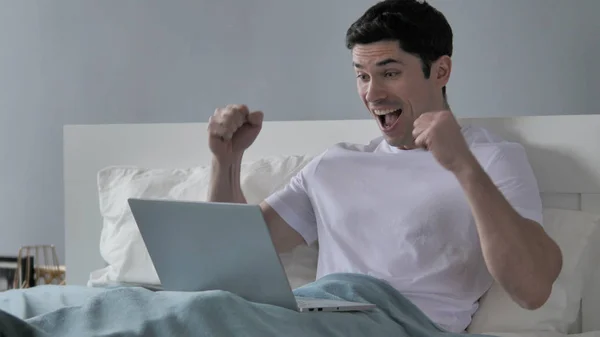 Giovane uomo che celebra il successo mentre lavora sul computer portatile a letto — Foto Stock