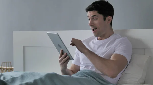 Mladý muž reagování na úspěch, používání tabletu v posteli — Stock fotografie