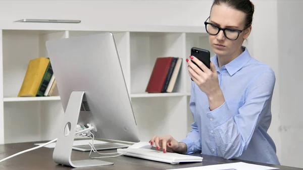 Mladá žena používání telefonu Smartphone při práci v počítači — Stock fotografie