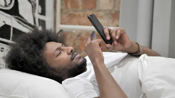 Afrikansk man med hjälp av smartphone, liggande i sängen — Stockfoto
