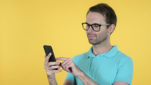 Junger Mann surft isoliert mit Smartphone auf gelbem Hintergrund — Stockvideo