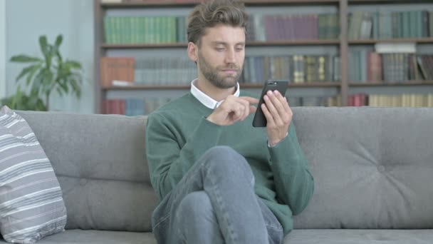 Молодой человек сидит на диване и просматривает свой смартфон — стоковое видео