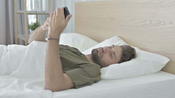 Geschokt jonge man praten op mobiele telefoon in bed — Stockvideo