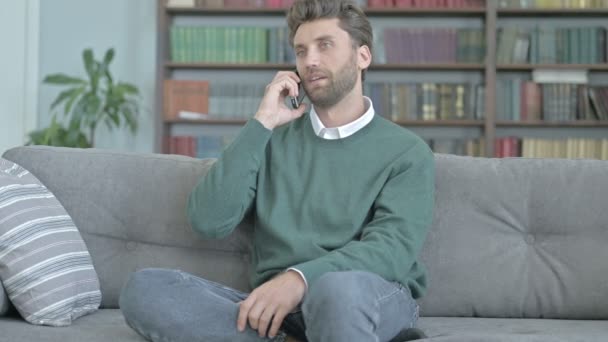 Jeune homme parlant au téléphone assis sur un canapé — Video