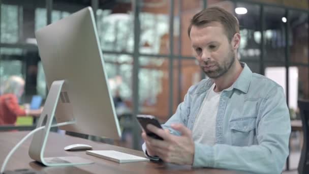 Creatieve jonge man met behulp van smartphone en computer op het werk — Stockvideo