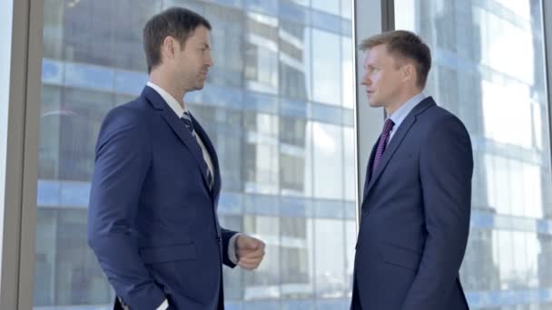 Dos hombres de negocios de mediana edad conversando y de pie contra la ventana de la oficina — Vídeo de stock