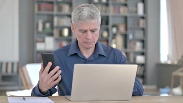 Seriöser Mann benutzt Smartphone bei der Arbeit am Laptop im Büro — Stockvideo