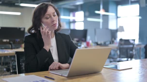 Alegre mujer de negocios de mediana edad que habla en el teléfono inteligente en el trabajo — Vídeo de stock