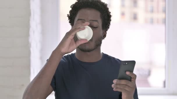 Retrato de un joven afroamericano bebiendo café y usando un teléfono inteligente — Vídeos de Stock