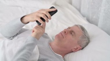 Yatakta akıllı telefon kullanan gevşemiş yaşlı adam. 