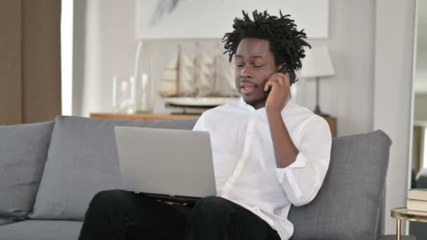 Afrikaanse Man met Laptop Praten op Smartphone in Home — Stockvideo