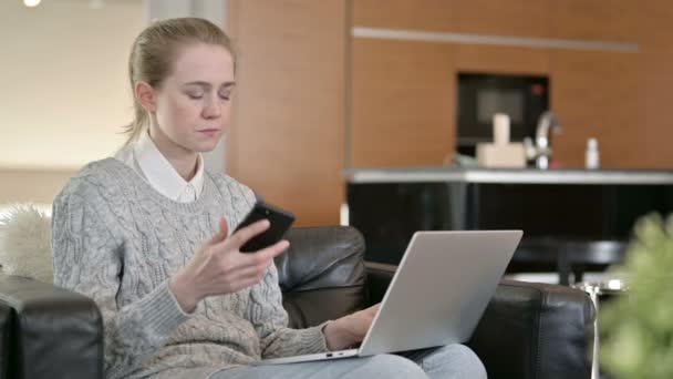 Молодая женщина с ноутбуком разговаривает на смартфоне дома — стоковое видео