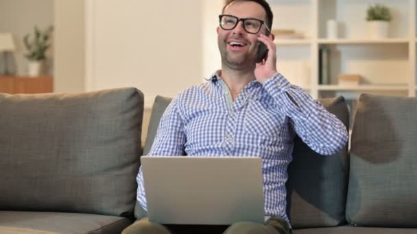 Весёлый человек с ноутбуком разговаривает на смартфоне — стоковое видео