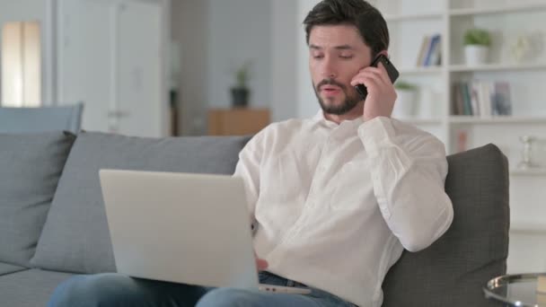 Junger Geschäftsmann mit Laptop zu Hause im Smartphone-Gespräch — Stockvideo