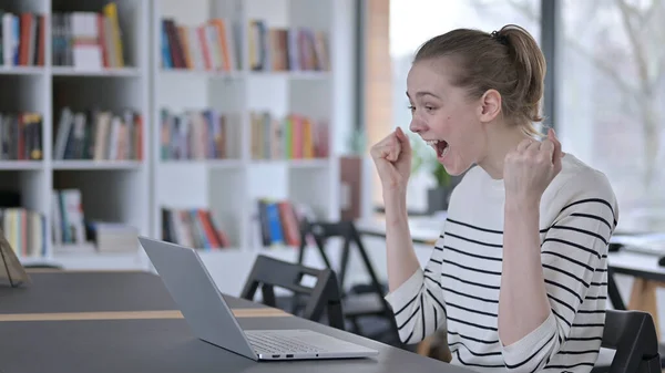 Pomyślna młoda kobieta świętująca na laptopie w bibliotece — Zdjęcie stockowe