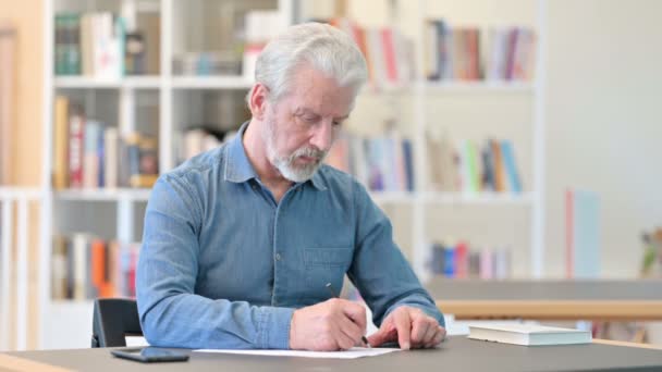 Vieil homme âgé concentré écrivant dans un carnet de notes à la bibliothèque — Video