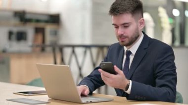 Ofiste akıllı telefon ve dizüstü bilgisayar kullanan genç bir iş adamı 
