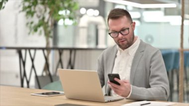Dizüstü bilgisayarlı sıradan bir genç ofiste akıllı telefondan konuşuyor. 