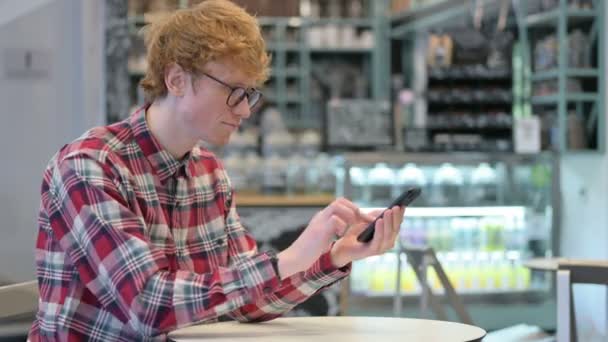 Joven pelirroja hombre celebrando el éxito en el teléfono inteligente en Café — Vídeos de Stock