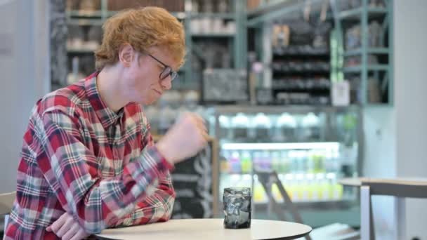 Genç kızıl saçlı adam su içiyor, kafede oturuyor., — Stok video